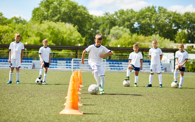 Portrait of boy in junior football team  leading ball between cones during  practice in field on sunny day with team in background