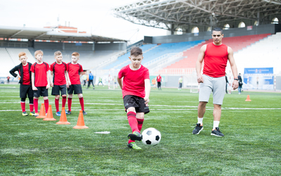 Full length portrait of junior football team practicing in stadium, focus on teenage boy leading ball with coach watching him intently, copy space